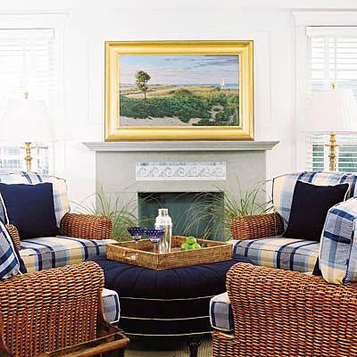 दो windows flank each side of the fireplace with a gray hearth. Also, white walls brighten up the space with yellow and blue large plaid cushions on wicker arm chairs.