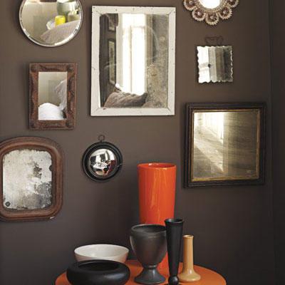 फंसाया mirrors of various sizes hung on a dark, brown wall with an orange circular table with various sized vases arranged on top