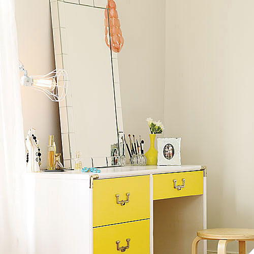  rectangular glass-framed mirror sits atop a yellow desk with a variety of perfume, make up brushes, photo frames and jewelry decorate the top of it