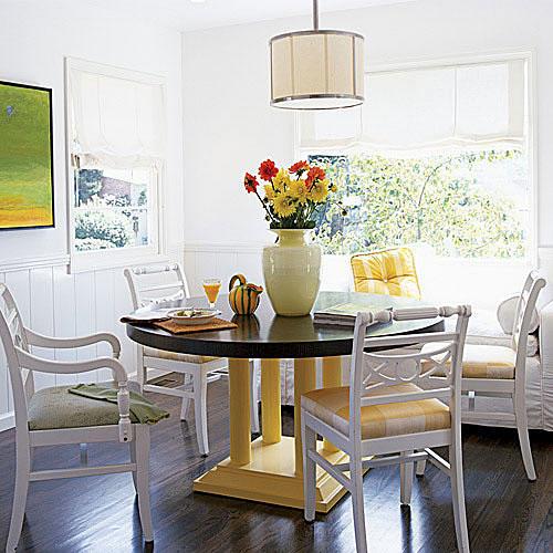  bright, white dining room with crisp, white cloth shades over the windows and a large circular wooden dining room table with pale, yellow legs and white chairs surround it