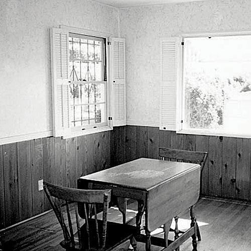  before photo of a dining room with pine boards up half of the wall that has windows with interior shutters. A small table with two chairs is positioned in the middle of the room.