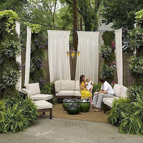 लंबा white drapes are hung around the driveway space with a loveseat with cotton cushions and side chairs arranged in a u-shape to create an outdoor room. Ferns in containers and other hanging flowers are hung throughout the space.